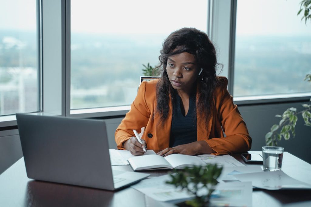 pexels-photo-5717313 A businesswoman in a modern office intently working on a laptop and writing in a notebook.