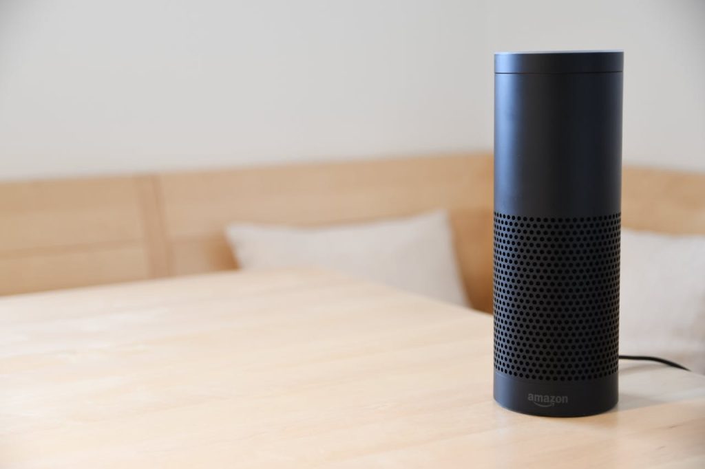 pexels-photo-977296 A black smart speaker resting on a light-colored wooden table in a cozy indoor setting.